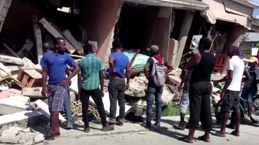 People stand in front of a collapsed building following an earthquake, in Les Cayes, Haiti, in this still image taken from a video obtained by Reuters on August 14, 2021.   REUTERS TV via REUTERS THIS IMAGE HAS BEEN SUPPLIED BY A THIRD PARTY. MANDATORY CREDIT.