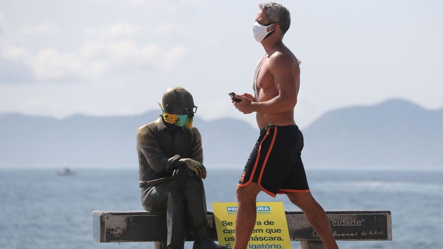 Um homem caminha ao lado da estátua do escritor brasileiro Carlos Drummond de Andrade que usa máscara protetora no primeiro dia de uso obrigatório de máscaras na cidade do Rio de Janeiro, em meio ao surto de doença por coronavírus (COVID-19), na praia de Copacabana